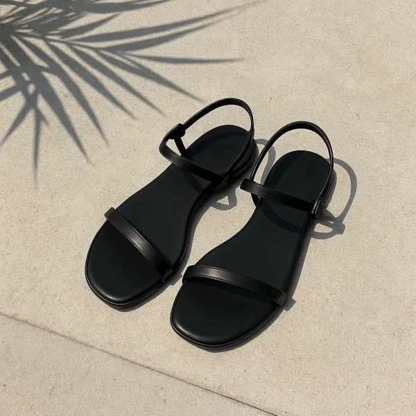 Μαύρα flat σανδάλια σε τσιμεντένια επιφάνεια με σκιά από καλοκαιρινά φυτά.